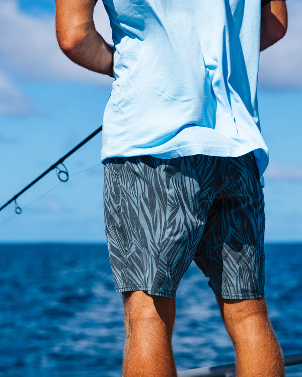 Wearing Salty Crew’s Transom 19" Overdye Short in Washed Green, a person stands on a boat fishing in the ocean under a blue sky. The photo, shot from behind, highlights the comfortable fit of these straight leg shorts.