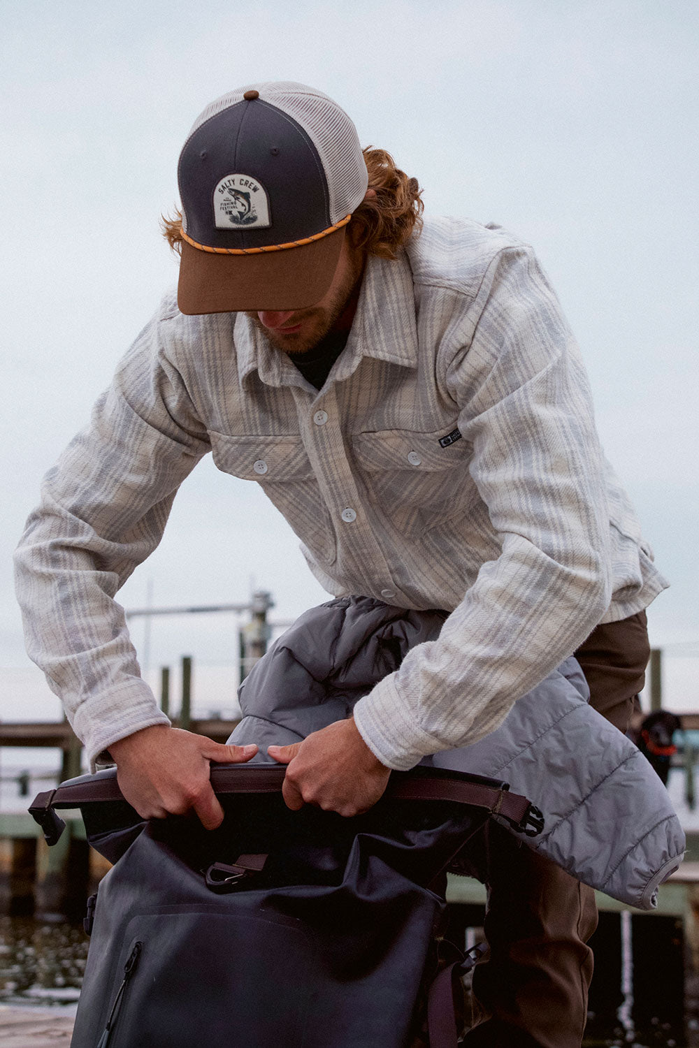 A person in a Salty Crew Windward Flannel in Off White and a cap is outdoors by a dock, packing a gray cotton heavyweight twill jacket with chest pockets into a black bag.