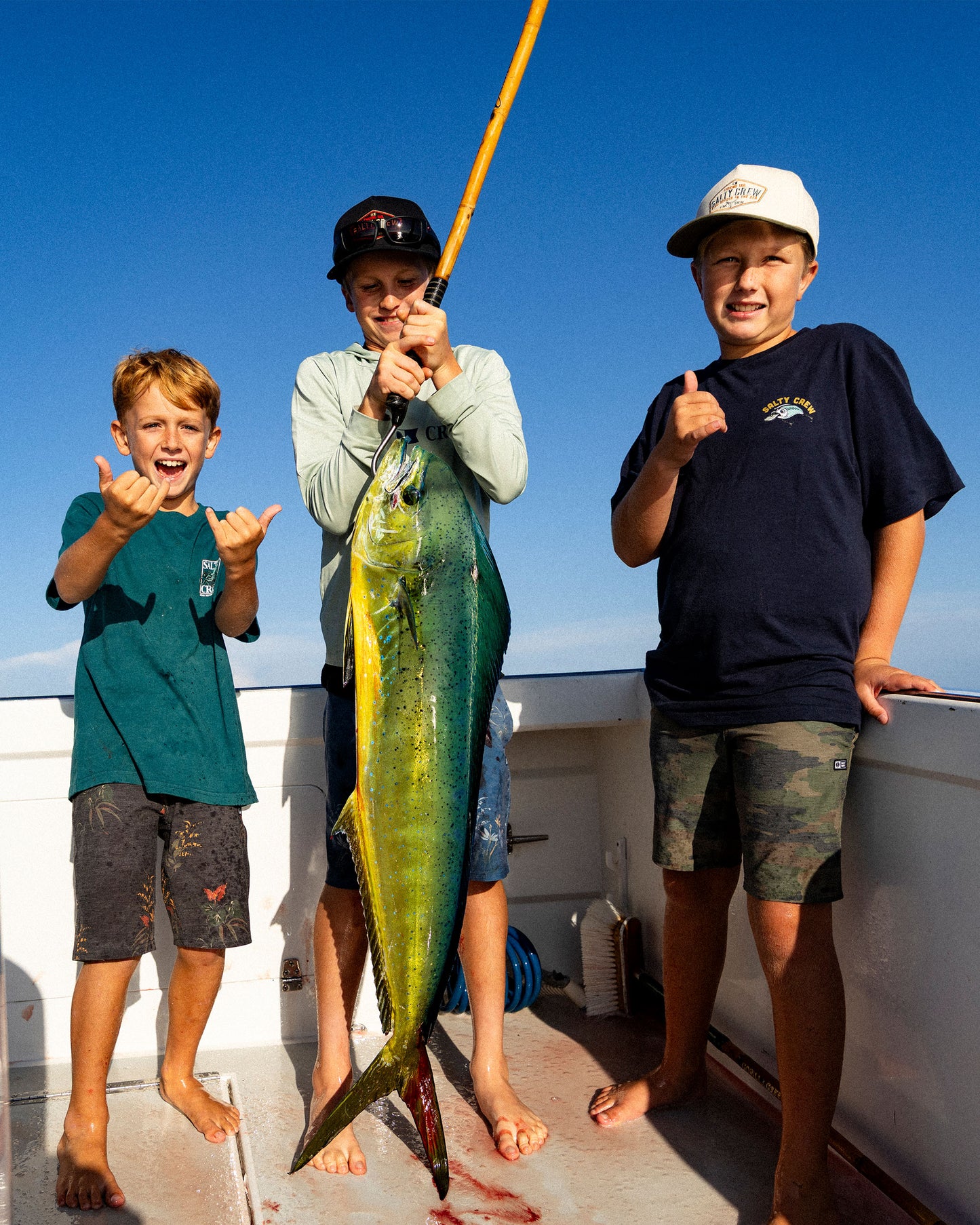 Three boys wearing Off Black Crew 17″ Boys Boardshorts by Salty Crew stand on a boat deck, the center one holding a large mahi-mahi fish on a line.