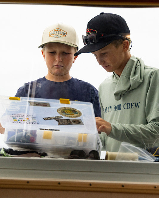 Two boys wearing black Tailfin Boys Snapback hats look down at an open plastic fishing tackle box.