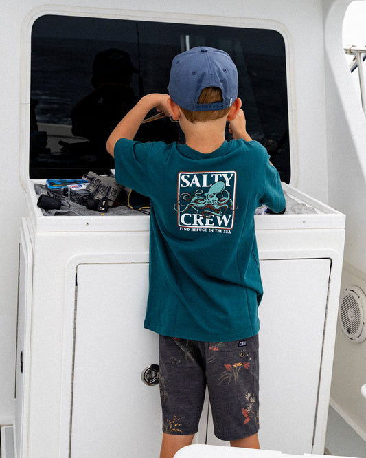 A toddler wearing a blue cap and a Salty Crew Ink Slinger Toddler Tee - Atlantic Blue with an octopus graphic on the back stands at a boat console.