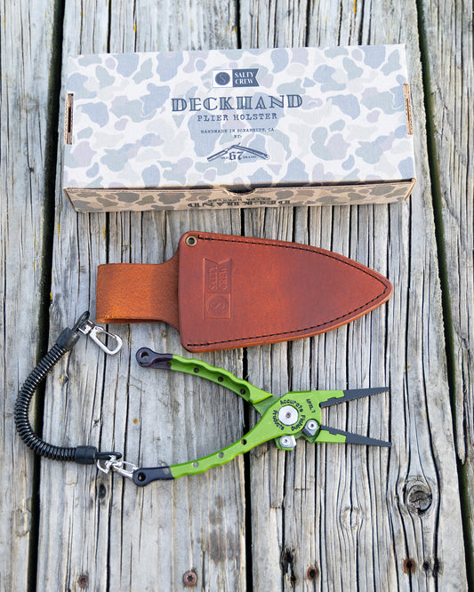 The Salty Crew Accurate x Salty Crew 7" Deckhand Plier with a green handle and black lanyard sits beside a brown leather holster and camo Deckhand Plier Holster box on weathered wood—essential fishing gear.