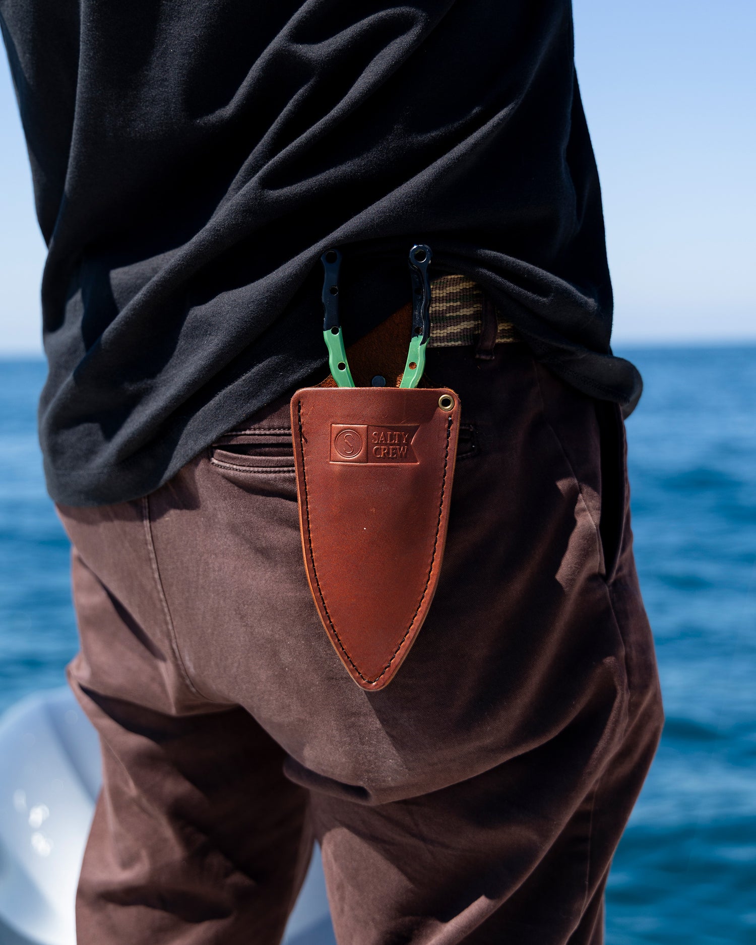 A person with brown pants and a black shirt stands by the ocean, carrying green-handled pliers in an Old 67 x Salty Crew Plier Holster by Salty Crew, attached to their fisherman's tool belt.