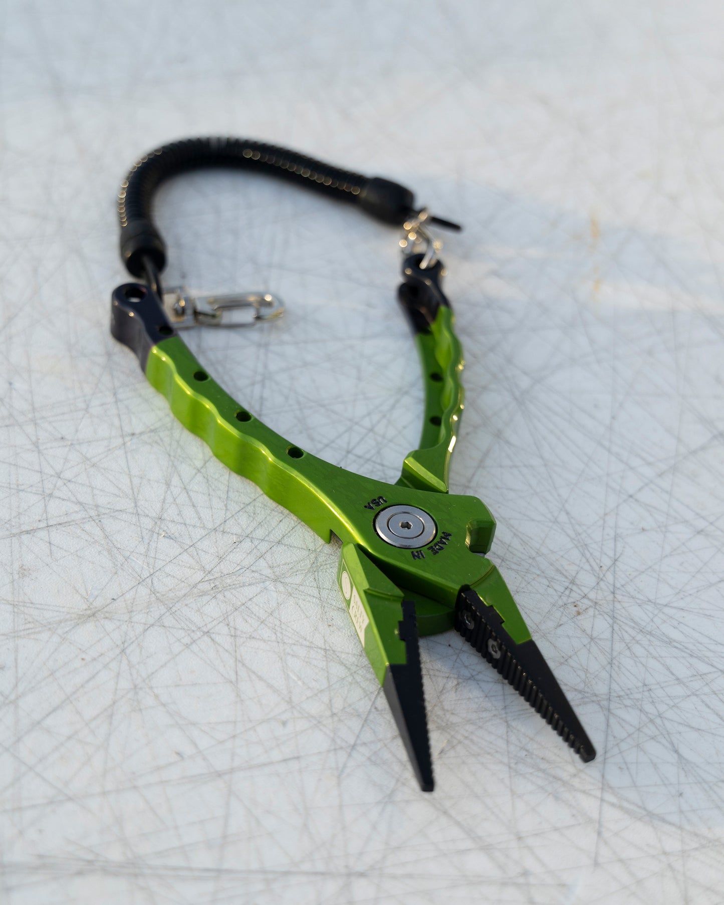 A Salty Crew Accurate x Salty Crew 7" Deckhand Plier in green with black jaws and a coiled lanyard sits on a scratched white surface.