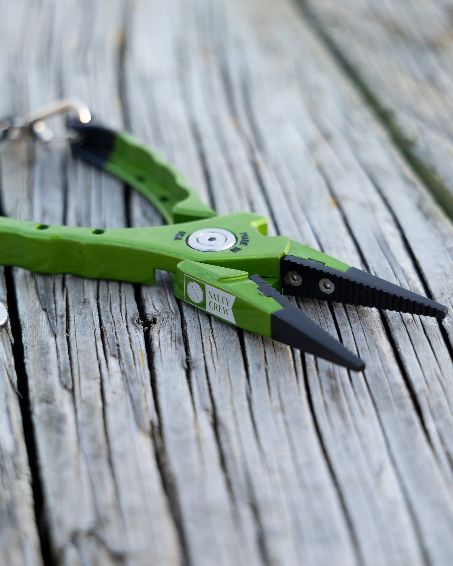 A pair of Salty Crew Accurate x Salty Crew 7" Deckhand Pliers in green and black with a keyring on one handle rests on weathered wooden boards.