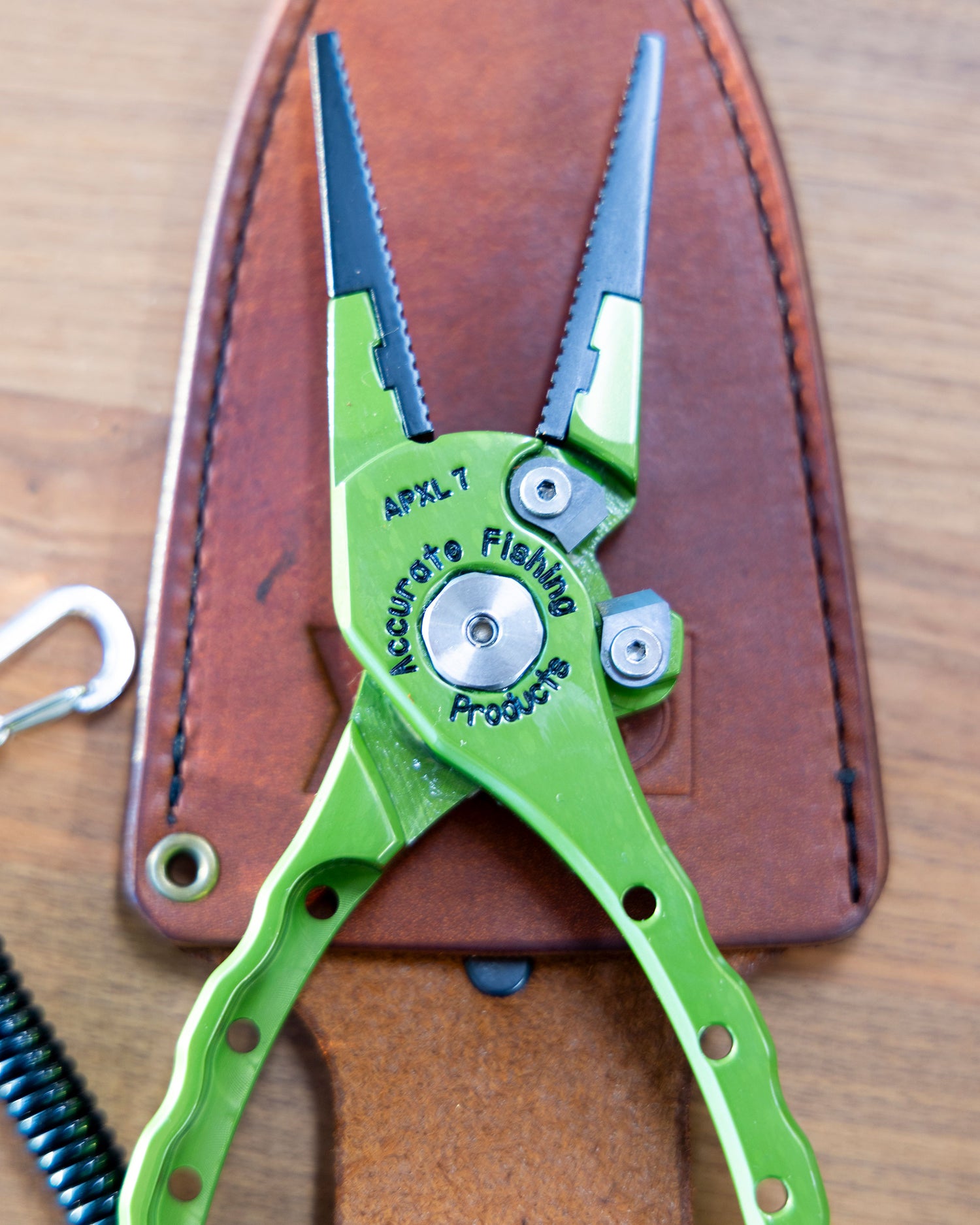 A pair of green Salty Crew Accurate x Salty Crew 7" Deckhand Pliers rests on a brown leather sheath atop a wooden surface, with a coiled black lanyard and metal clip partially visible on the left.