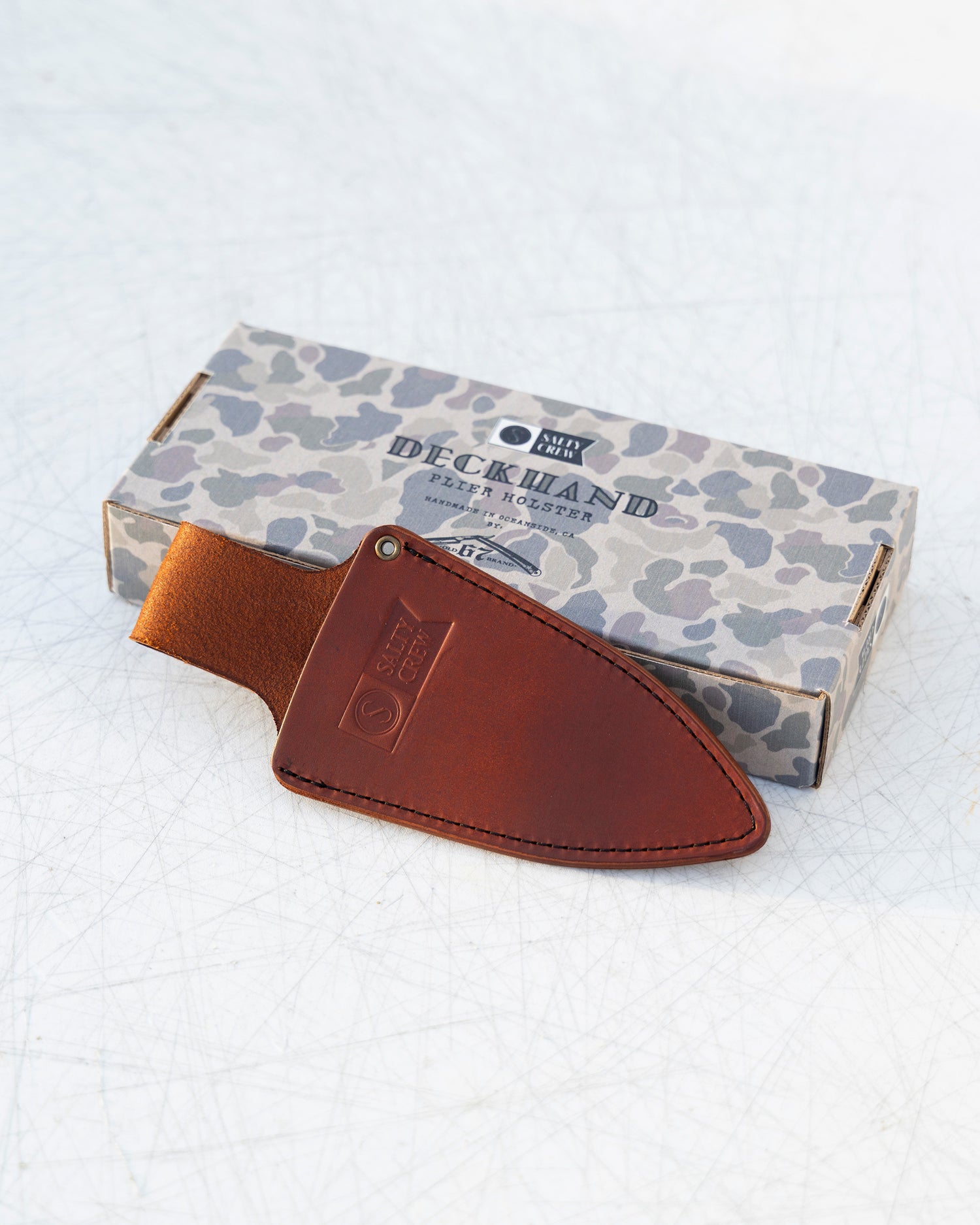 The Old 67 x Salty Crew Plier Holster, a handcrafted sheath made from premium veg tan leather, is shown in front of a camouflage-patterned Salty Crew box on a white textured surface.