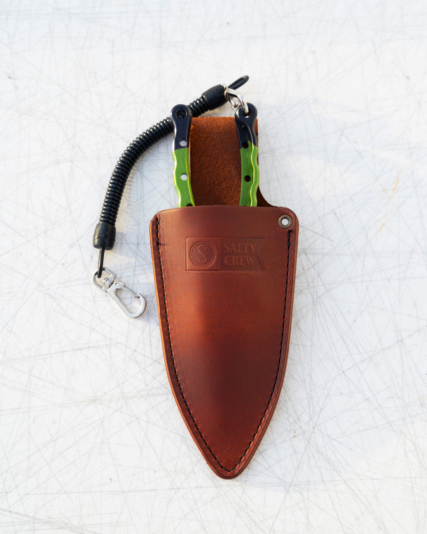 The Old 67 x Salty Crew Plier Holster by Salty Crew features green-handled pliers with a coiled lanyard and clip in a handcrafted, logo-embossed leather sheath—perfect for any angler’s tool belt.