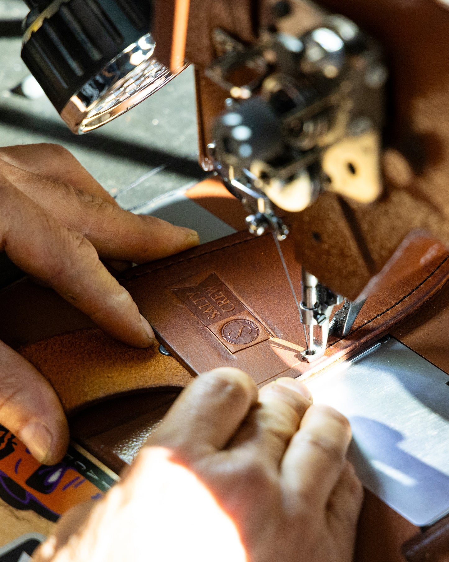 Close-up of hands stitching premium veg tan leather stamped with HAND MADE for the Old 67 x Salty Crew Plier Holster by Salty Crew. Bright light highlights expert craftsmanship at the sewing machine.