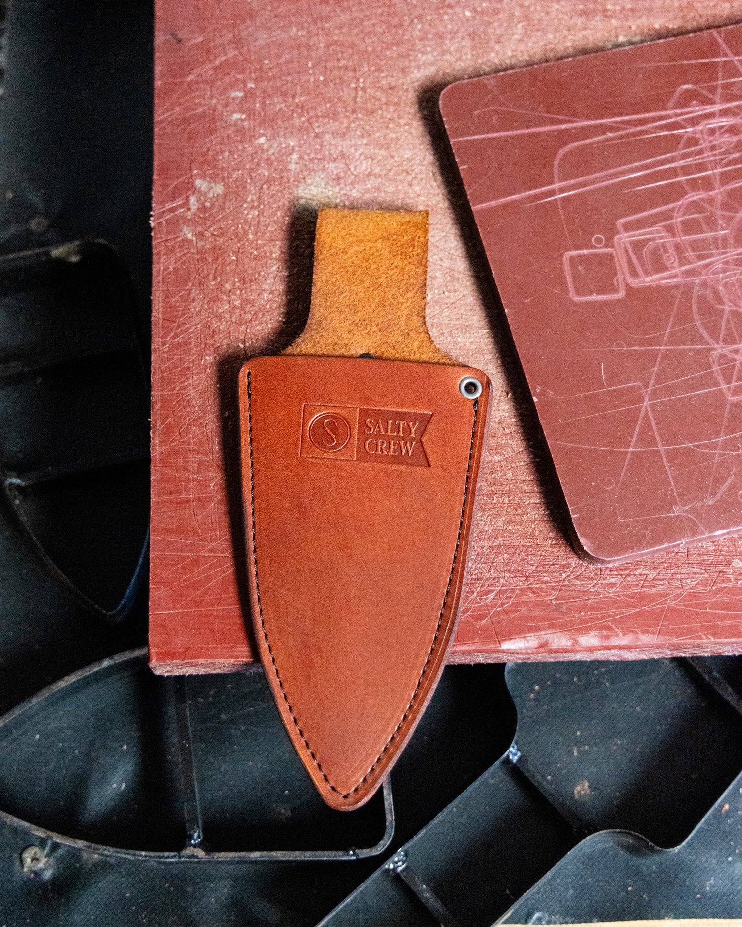 The Old 67 x Salty Crew Plier Holster, crafted from premium veg tan leather and stamped with the Salty Crew logo, lies on a red textured surface beside a scratched brown rectangular item with black metal partially visible.