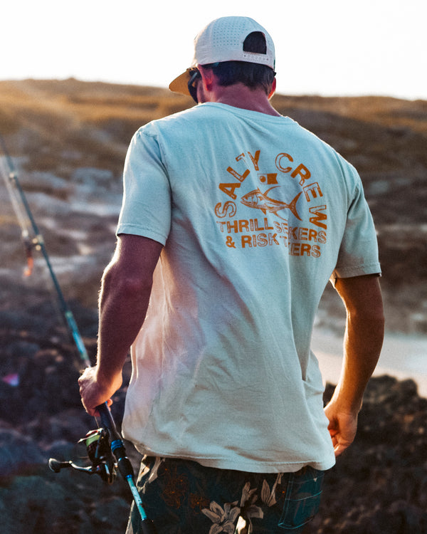 A man holding a fishing rod walks over rocky terrain. He wears a white cap and a beige T-shirt with the words “Salty Crew Thrill Seekers & Risk Takers” printed on the back, along with a fish graphic.