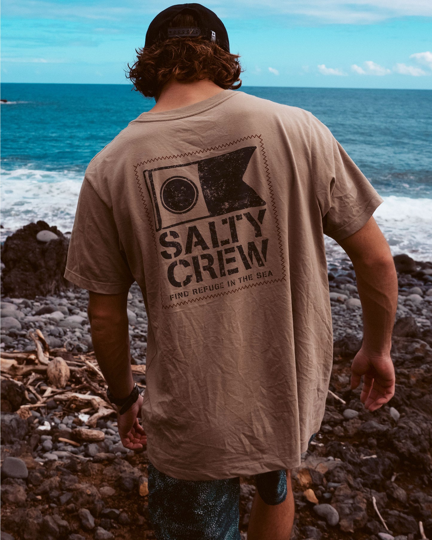 Wearing a black cap and a tan Salty Crew Stitched Tee - Twig, a person with wavy brown hair stands on rocky shore facing the ocean under a partly cloudy sky.