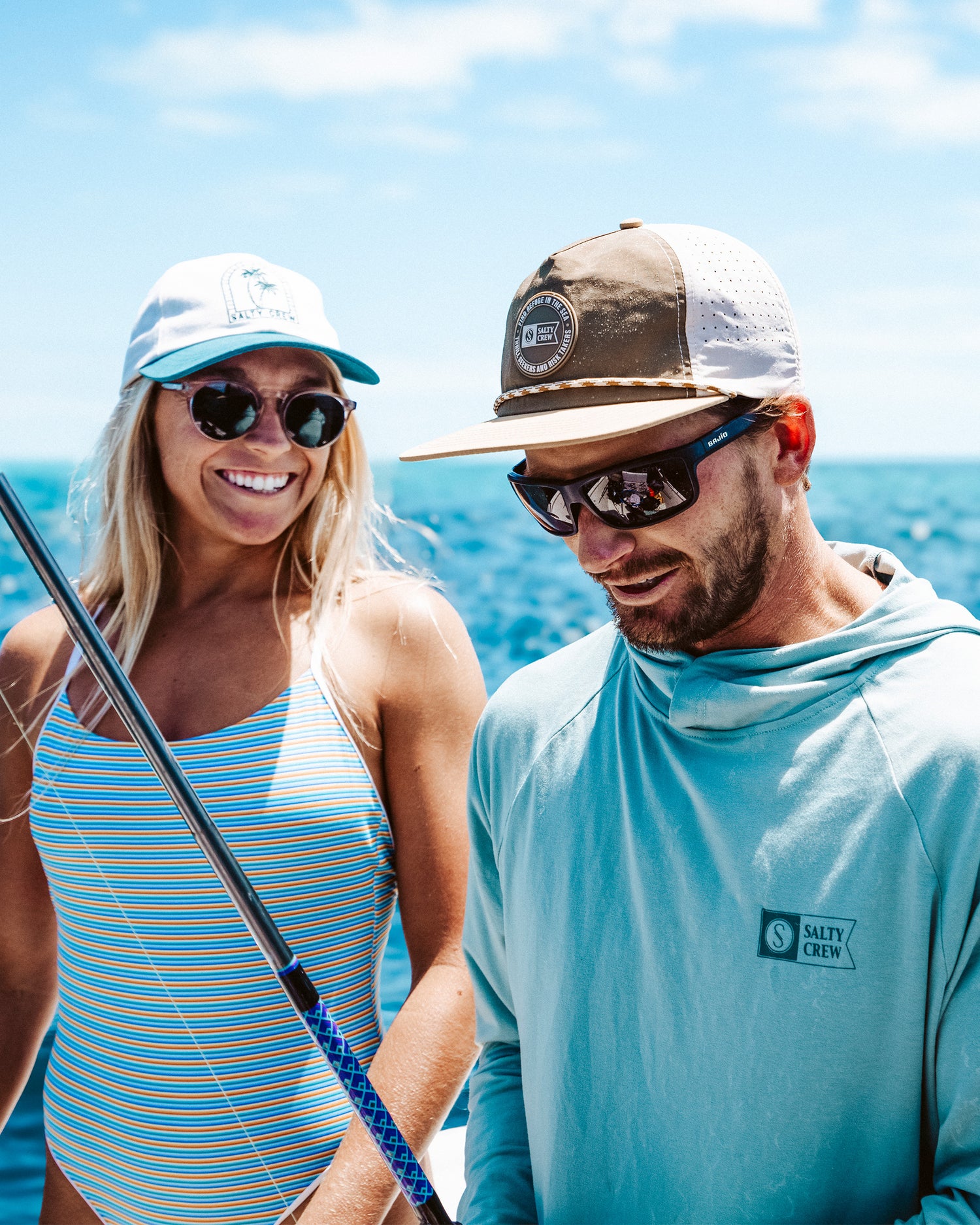 A woman in a striped swimsuit and a man wearing a Salty Crew Topshot Tech Snapback - Olive/Gold and a light hoodie smile together on a boat, with the ocean and blue sky behind them.