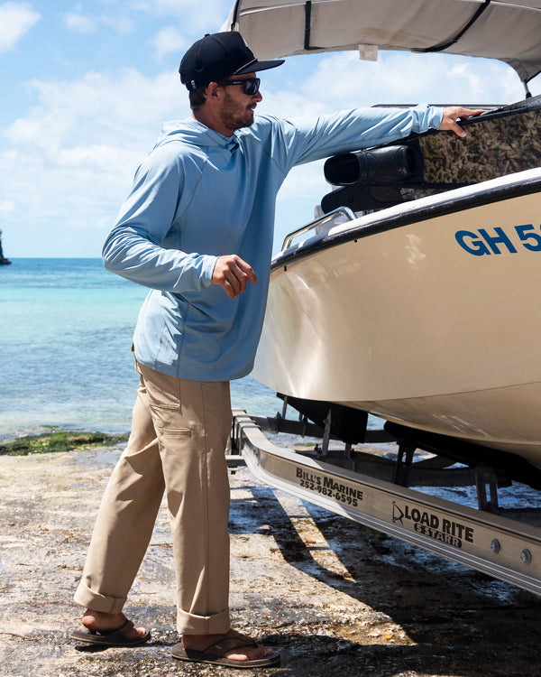 A man wearing Salty Crew Transom Tech Pants in khaki, a hat, sunglasses, and a light blue quick-dry shirt stands by a boat on a trailer at the shore, reaching toward the boat with the ocean behind him.