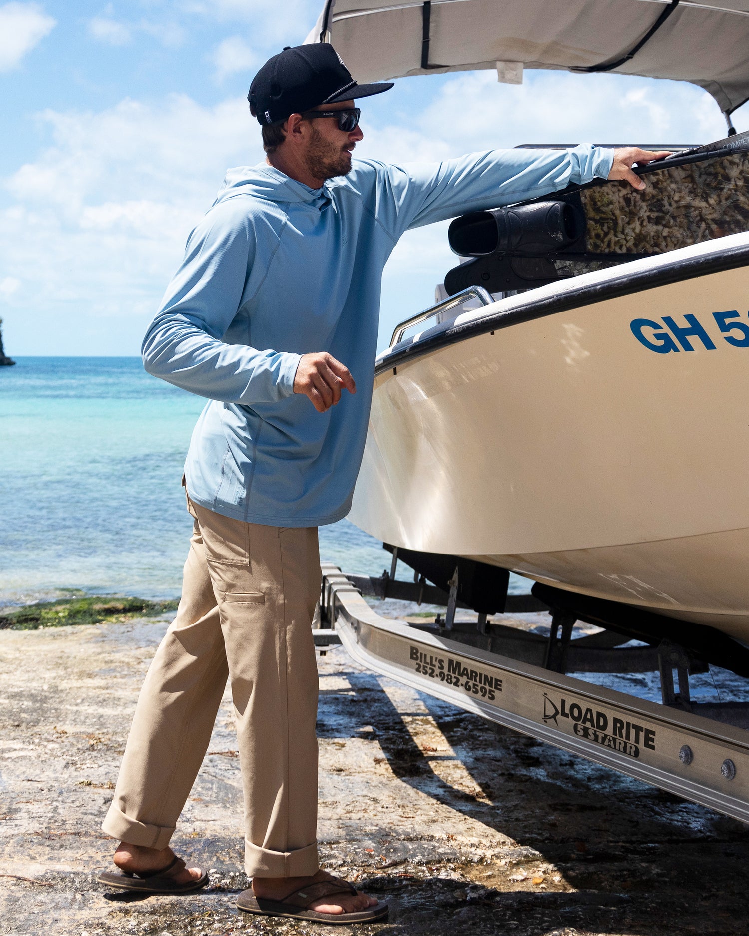 A man wearing Salty Crew Transom Tech Pants in khaki, a hat, sunglasses, and a light blue quick-dry shirt stands by a boat on a trailer at the shore, reaching toward the boat with the ocean behind him.