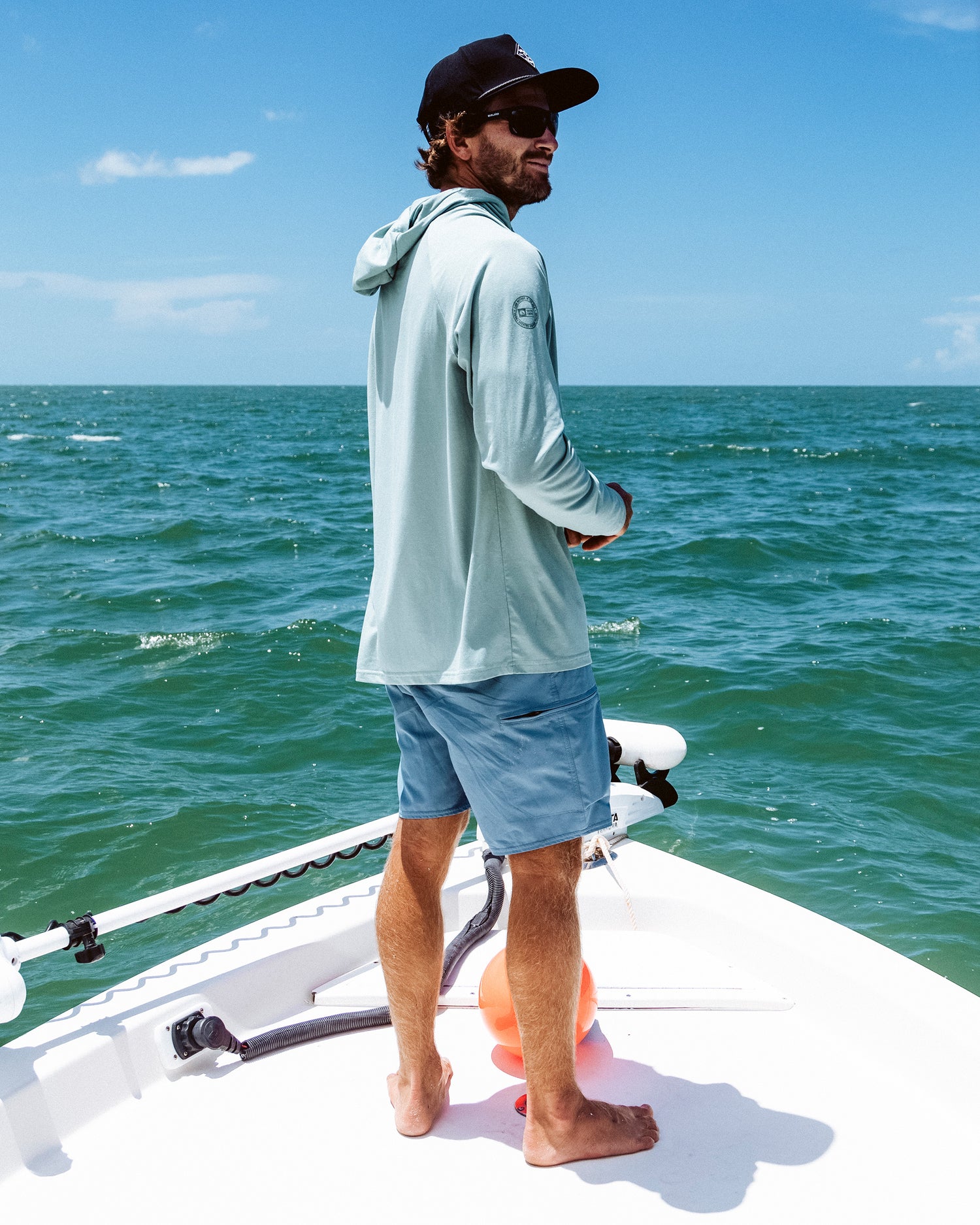 A man in a light hooded shirt, Salty Crew Transom 19" Tech Short in Pilot Blue, and a cap stands barefoot on a boat, gazing at the ocean under a sunny sky with the sea and blue horizon in the background.