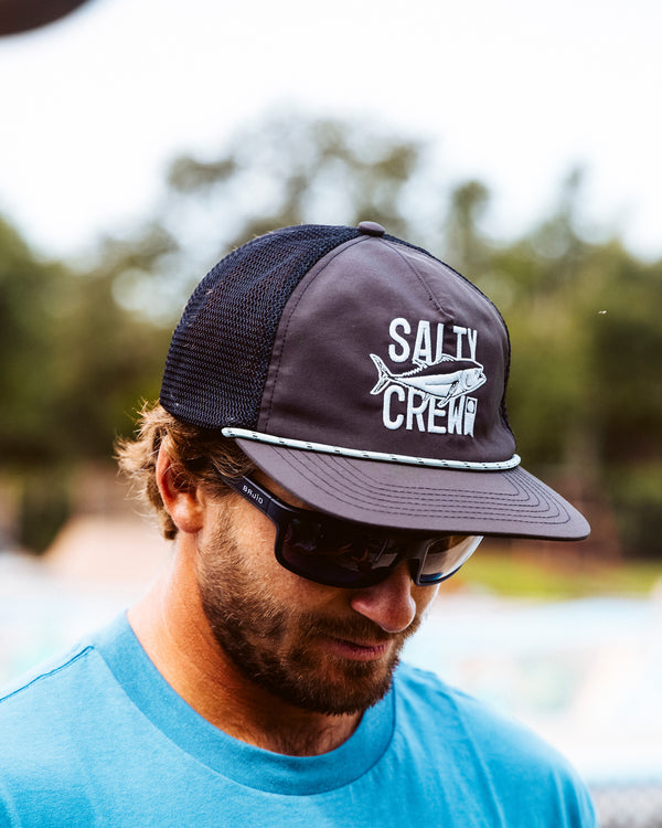 A man wearing Salty Crew's Tried And True Trucker - Phantom, a black mesh-back snapback with a fish logo, black sunglasses, and a blue shirt looks down with blurred greenery in the background.