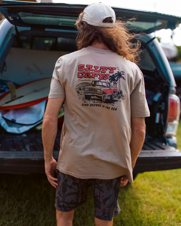 A person with long hair, wearing the Salty Crew Truck Stop Classic Tee - Twig made from soft spun 100% cotton and dark shorts, stands by an open vehicle trunk filled with surfboards on grass.