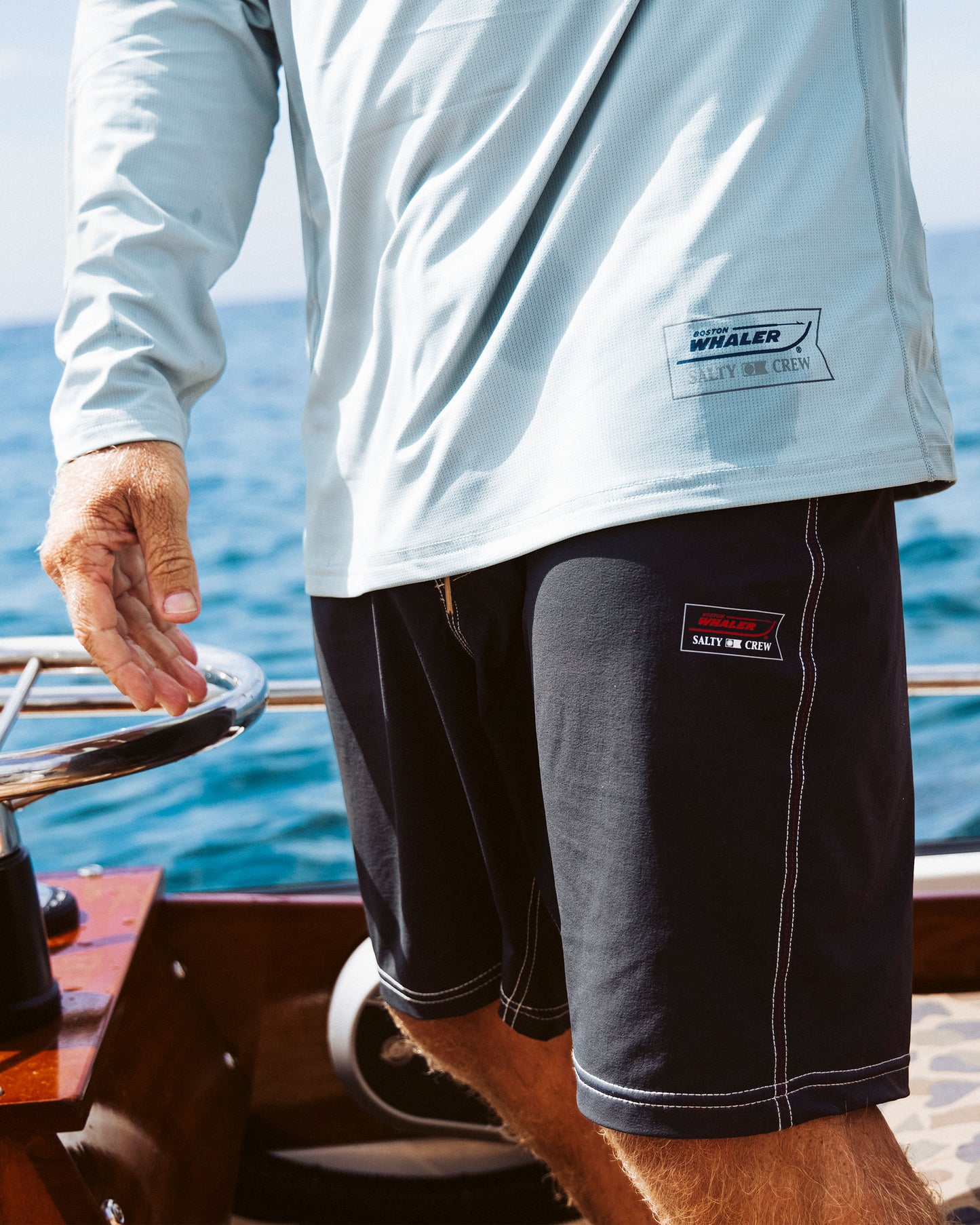 A person wearing a light blue long-sleeve shirt and Salty Crew Whaler 18.5" Boardshort - Black stands on a boat, steering with the ocean in the background. The shirt and shorts feature boating-themed patches.