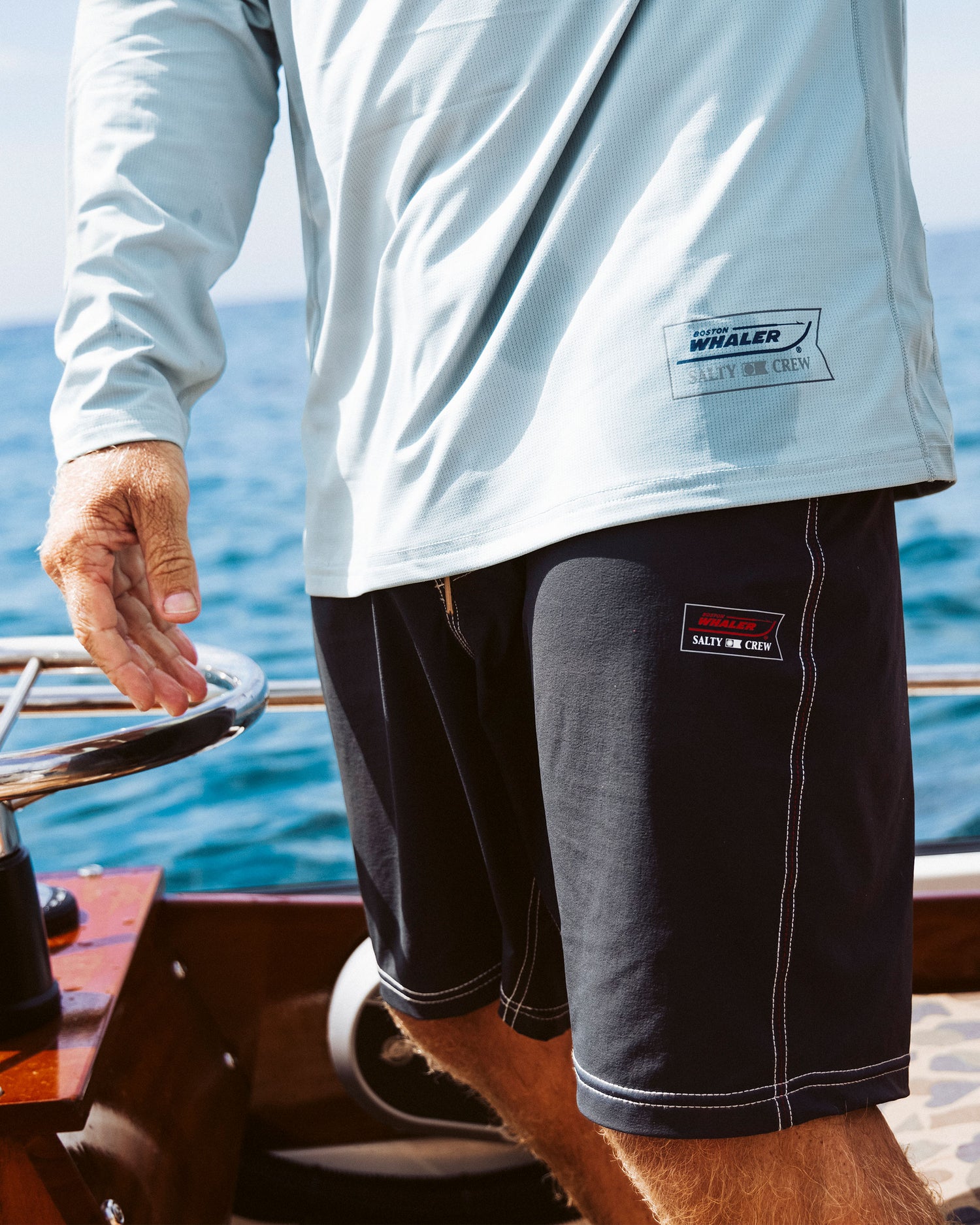 A person wearing a light blue long-sleeve shirt and Salty Crew Whaler 18.5" Boardshort - Black stands on a boat, steering with the ocean in the background. The shirt and shorts feature boating-themed patches.