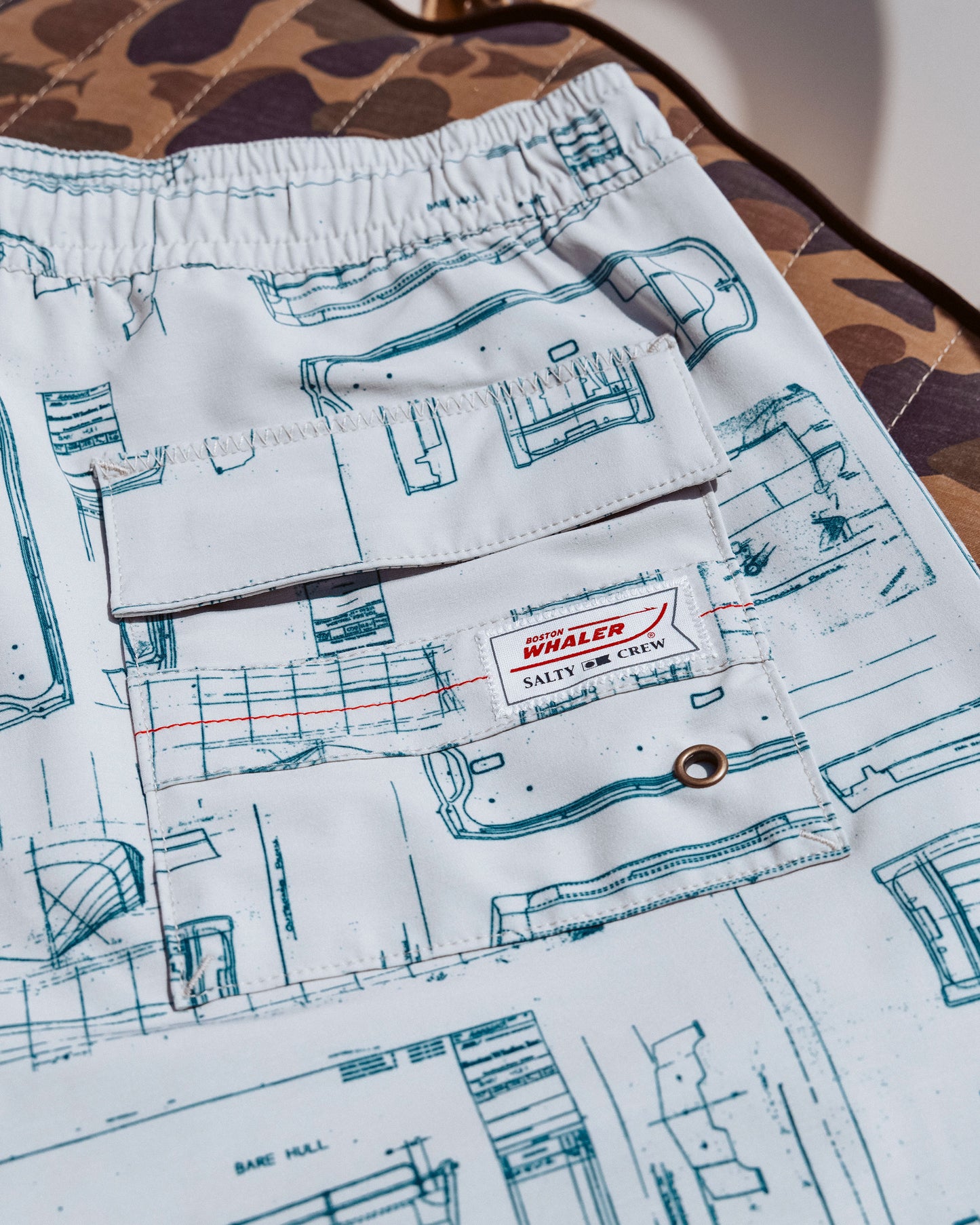 Close-up of Salty Crew Whaler Volley 18" shorts in Bone, crafted from quick-dry recycled polyester with a blue blueprint pattern, flap pocket with "WHALER SALTY CREW" label, and metal grommet on brown camo background.