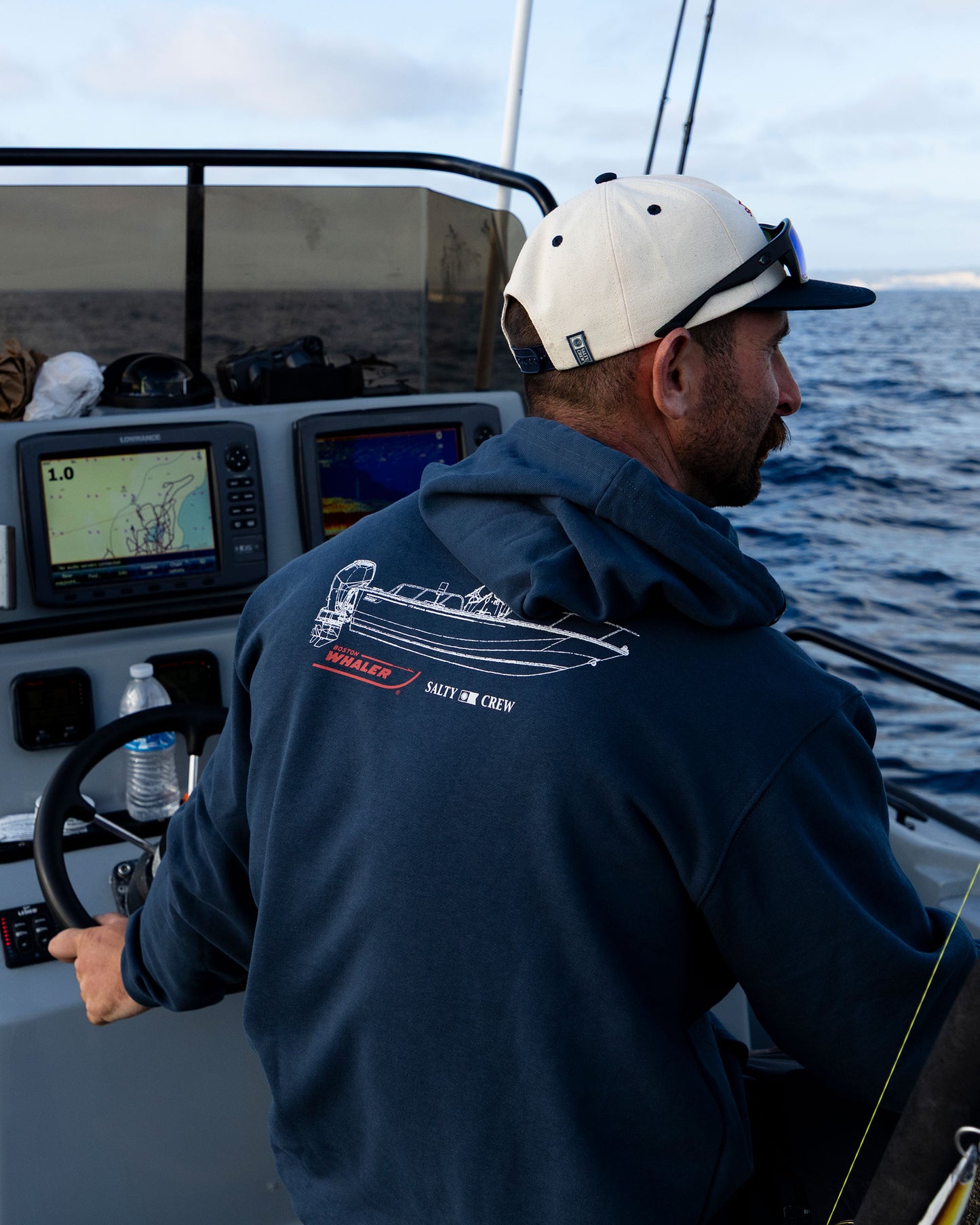 A man steers a boat wearing the Salty Crew Whaler Schematic Fleece in navy, featuring a screenprinted boat graphic and SALTY SIR CREW text. Boat controls, navigation screens, fishing rods, and his white cap are visible.