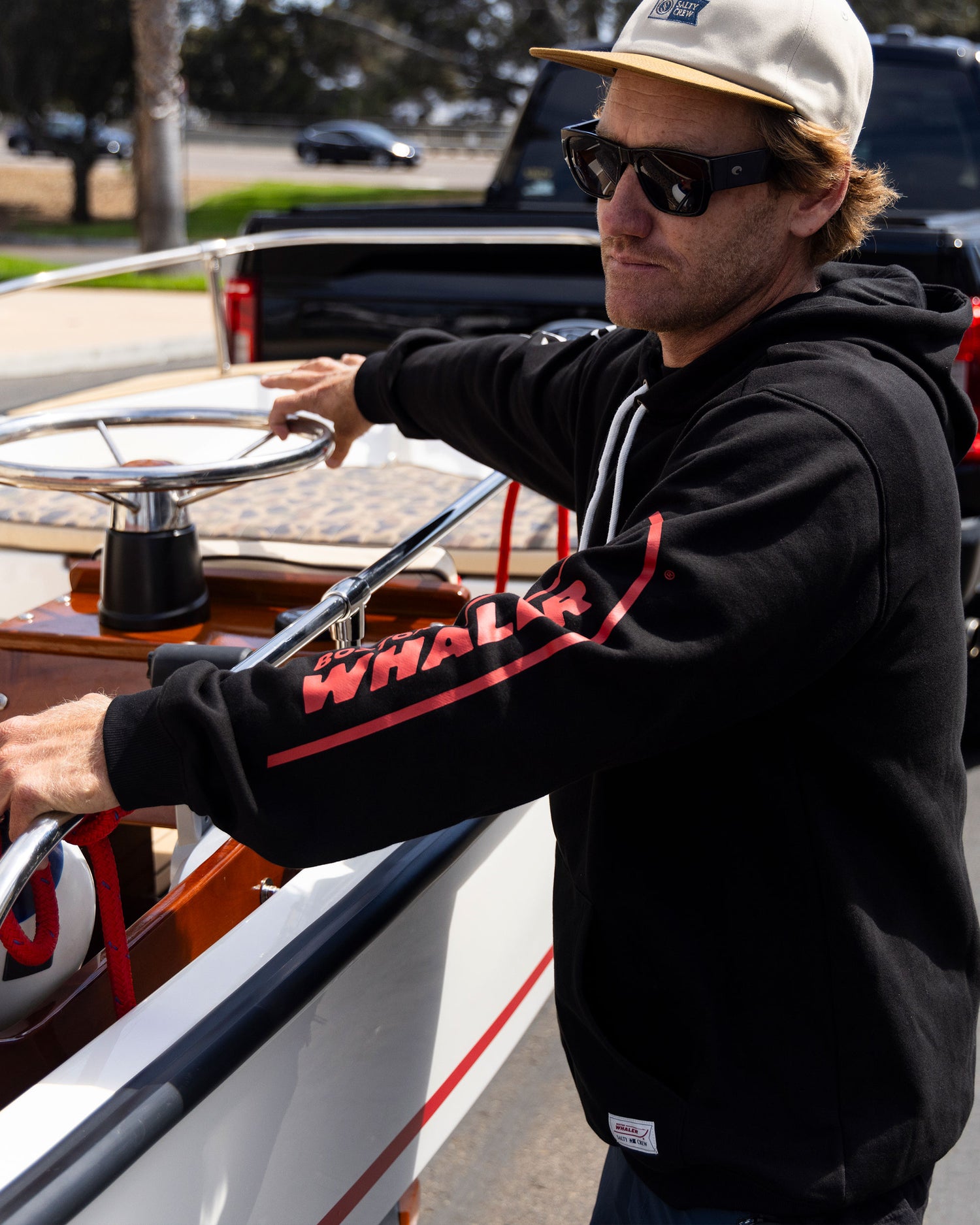 A man in Salty Crew’s Whaler Icon Fleece - Black, sunglasses, and a tan cap stands by a boat, gripping the steering wheel on a sunny day. The black cotton-poly hoodie features screenprinted "WHALER" on the sleeves.