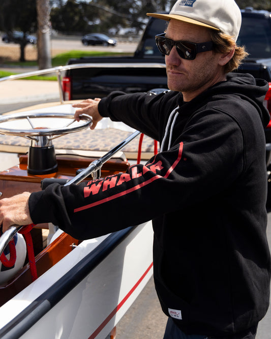 A man in Salty Crew’s Whaler Icon Fleece - Black, sunglasses, and a tan cap stands by a boat, gripping the steering wheel on a sunny day. The black cotton-poly hoodie features screenprinted "WHALER" on the sleeves.