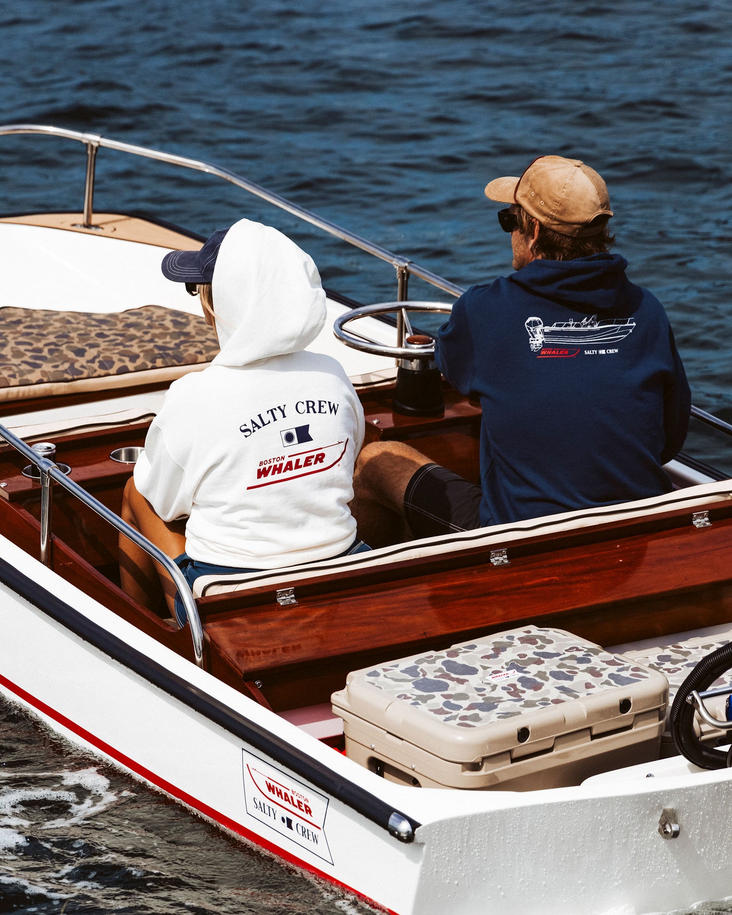Two people sit in a small boat with wood accents and a camouflage cooler. One wears the Whaler Hoodie - Off White, the other a Salty Crew sweatshirt—both featuring boating-themed graphics.