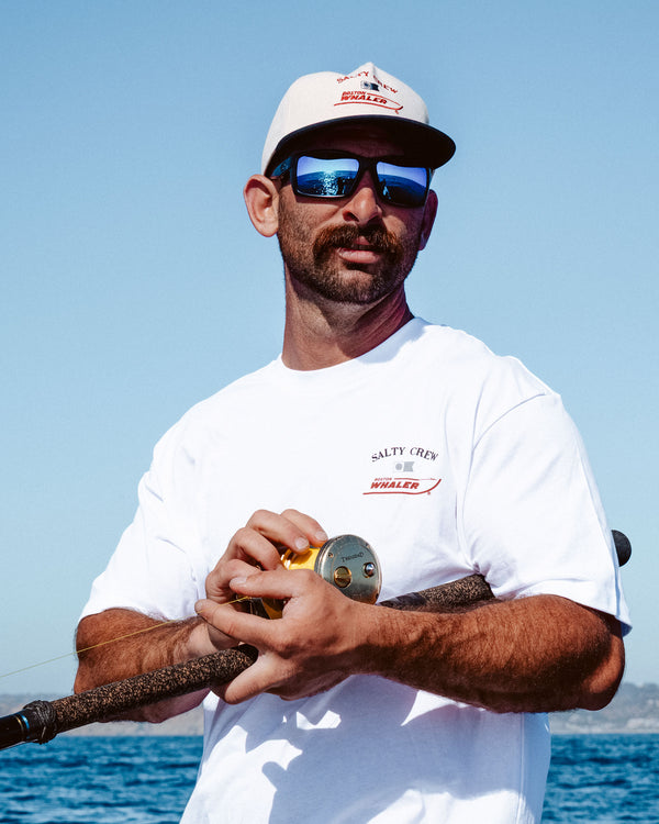 A man in a white T-shirt, sunglasses, and the Salty Crew Whaler Snapback - Off White stands outdoors with a fishing rod, blue sky, and sea in the background.