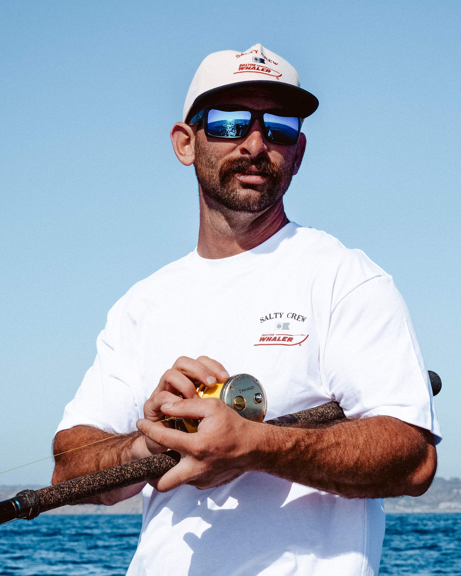 A man in a white T-shirt, sunglasses, and the Salty Crew Whaler Snapback - Off White stands outdoors with a fishing rod, blue sky, and sea in the background.