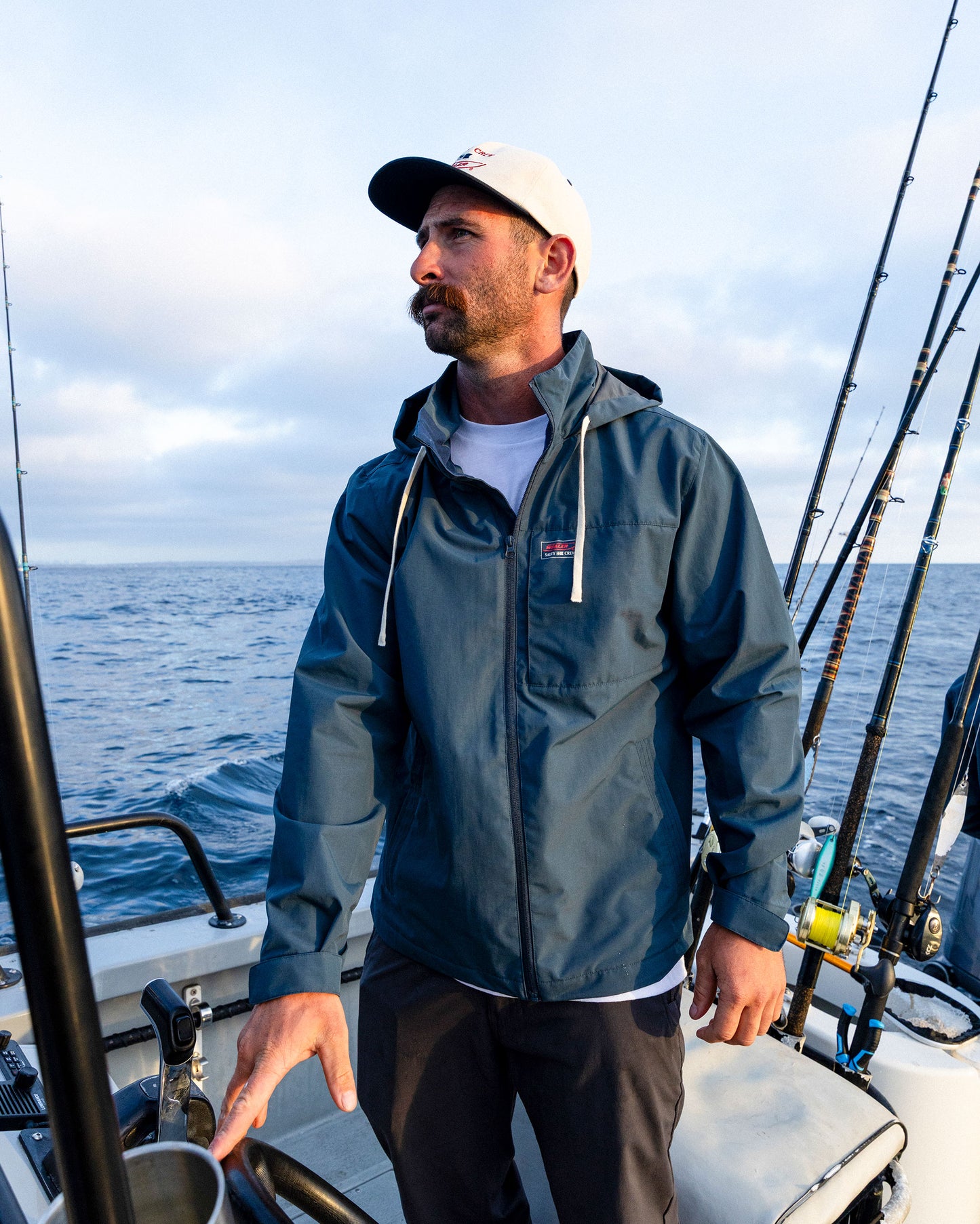 A man wearing the Salty Crew Whaler Windbreaker in navy and a white cap stands on a boat amid fishing rods, with the ocean and a cloudy sky behind him.