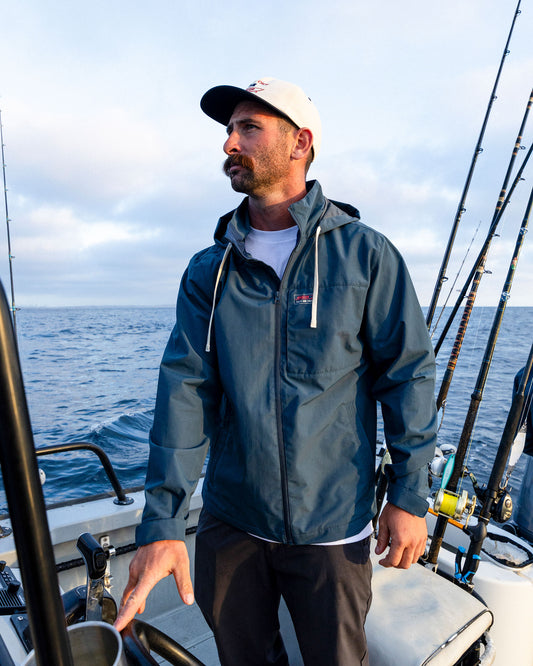 A man wearing the Salty Crew Whaler Windbreaker in navy and a white cap stands on a boat amid fishing rods, with the ocean and a cloudy sky behind him.