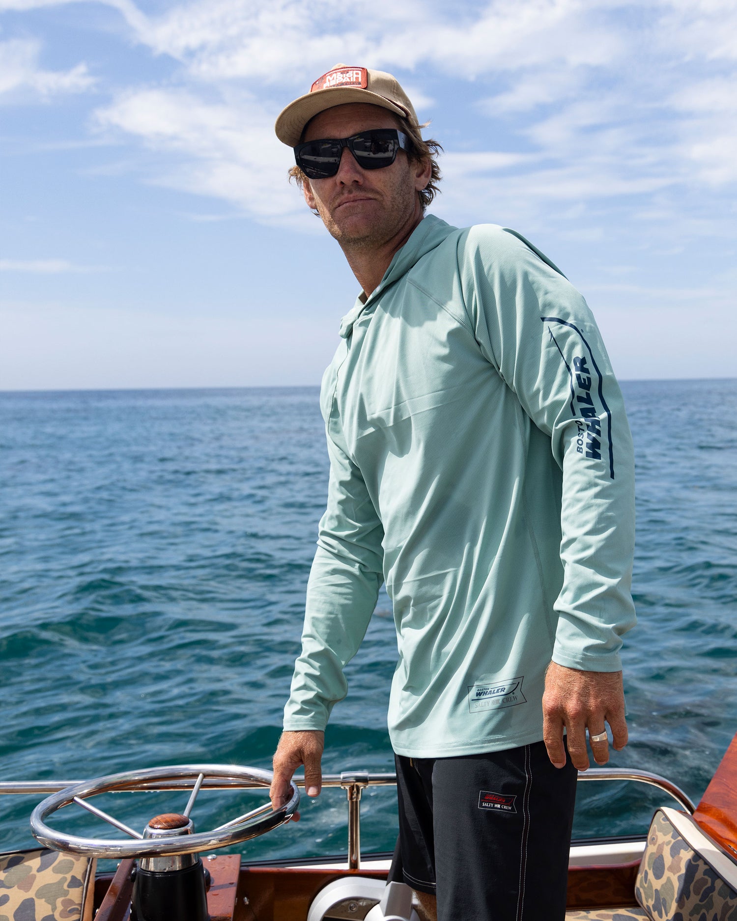 A man in sunglasses and a cap stands at the wheel of a boat, wearing the Salty Crew Whaler Apex Sunshirt - Mackerel, a light blue UPF 50+ long-sleeve, with blue ocean water and partly cloudy sky in the background.