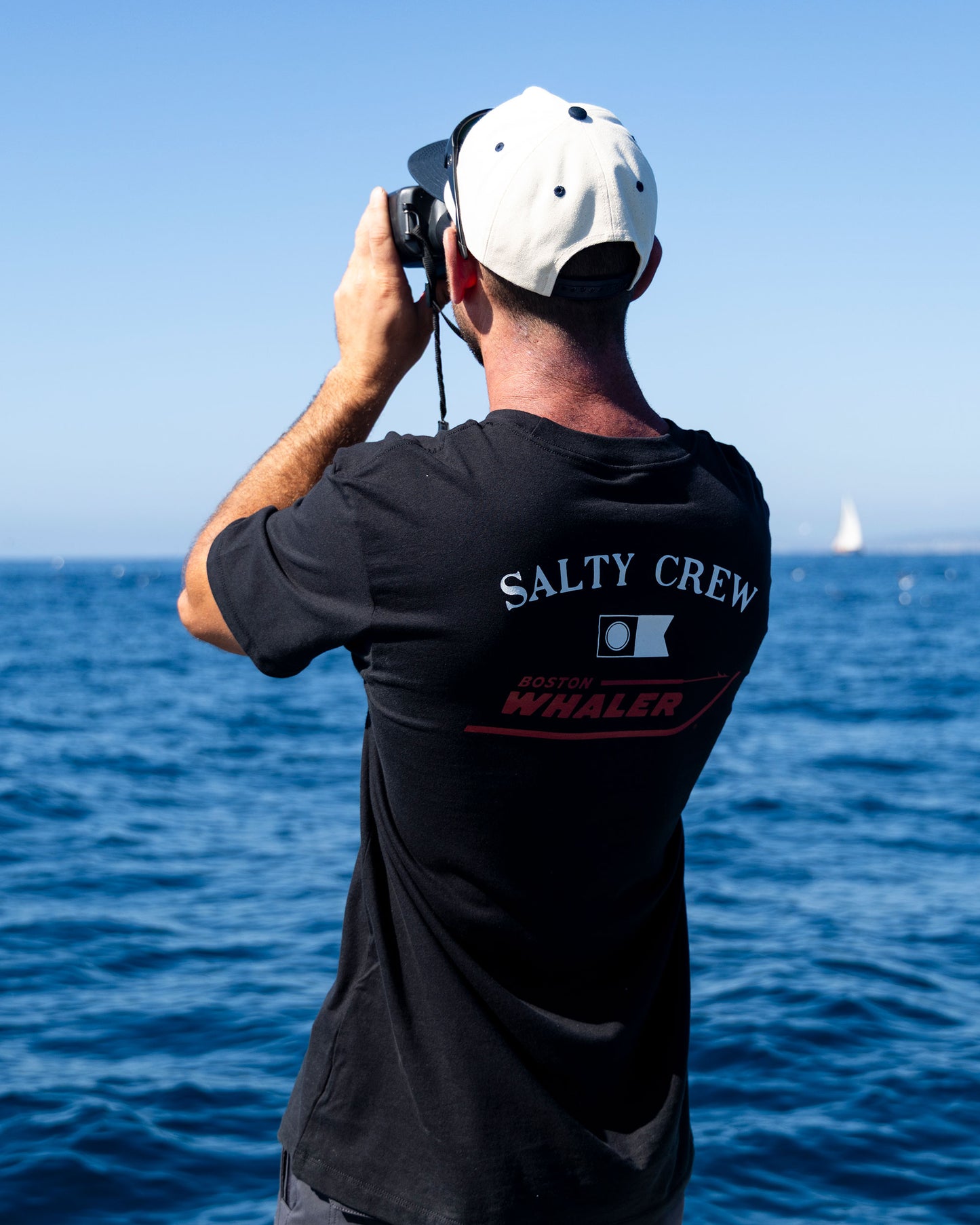 Wearing a black Salty Crew Whaler Icon Crew Tee made from 100% combed ringspun cotton and a white cap, a person stands by the ocean, gazing through binoculars on a sunny day as a sailboat appears in the distance.