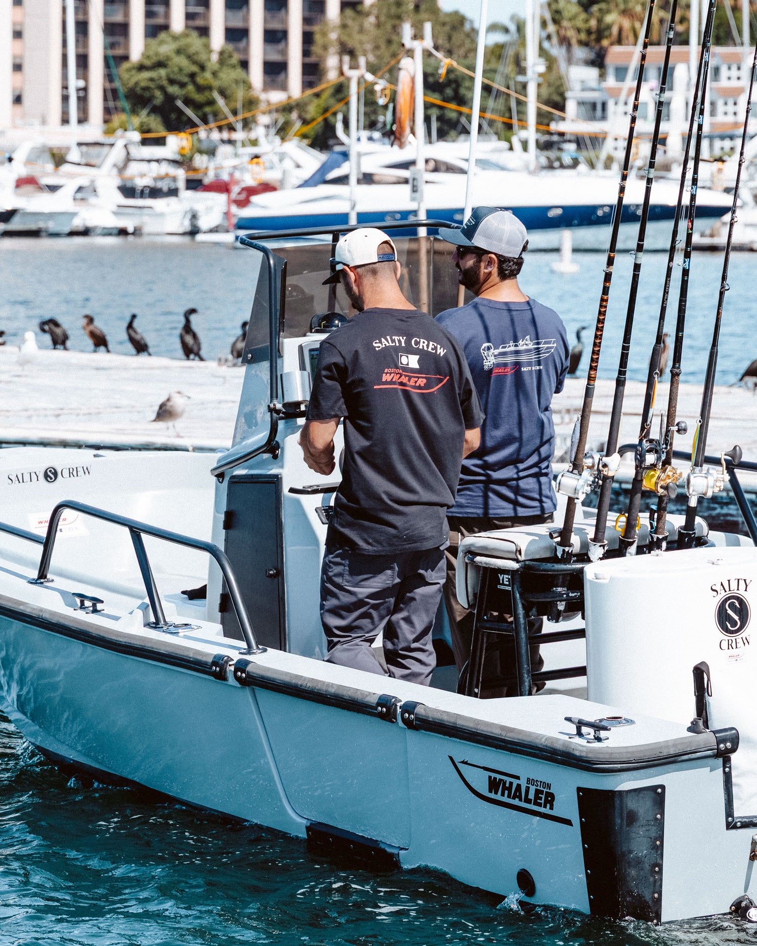 Two men stand on a small white fishing boat near a marina, wearing Salty Crew Whaler Schematic Crew Tees in navy. Surrounded by fishing rods, they face away from the camera as birds gather on the dock nearby.