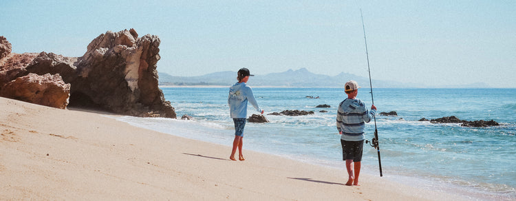 Two boys in casual beach clothes fish along a sandy shoreline, one holding a fishing rod and the other casting, with rocky formations and calm blue ocean waves in the background under a clear sky.