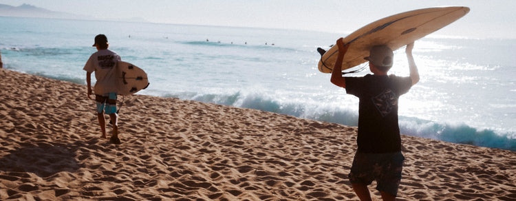 Two people walk along a sandy beach toward the ocean, each carrying a surfboard, with gentle waves and distant surfers visible in the background under a bright sky.