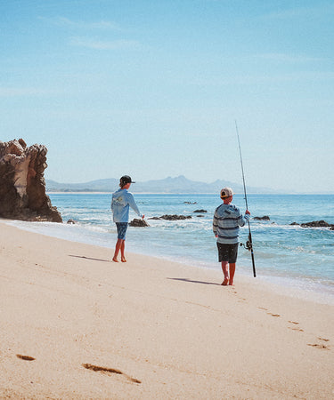 Two children walk on a sandy beach near the water; one holds a fishing rod. Rocky formations and gentle waves are visible, with mountains in the distant background under a clear blue sky.