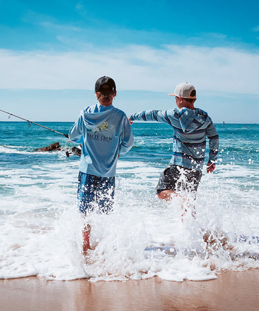Two boys stand at the edge of the beach, one fishing and the other kicking up water as waves splash around them under a blue sky.