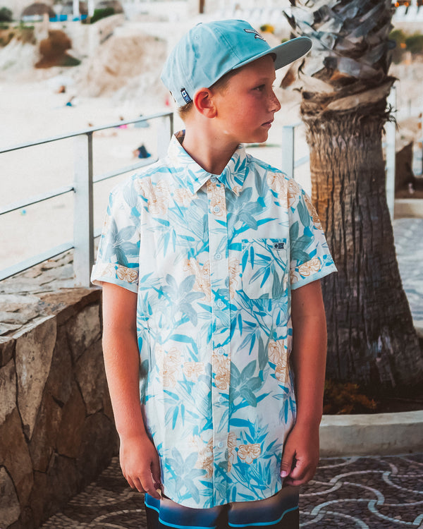 A young boy in a pale blue cap and a floral-patterned short-sleeve shirt stands outdoors by a palm tree, looking to the side near a beachside walkway with people and sand visible in the background.