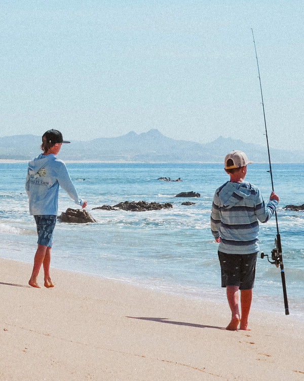 Two boys stand barefoot on a sandy beach; one holds a fishing rod, while the other looks toward the ocean. Rocky formations and distant mountains are visible under a clear blue sky.