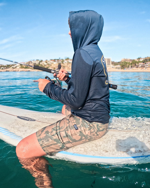 A person wearing a navy hooded shirt and camouflage shorts is sitting on a paddleboard, fishing on clear blue water with buildings visible on the shore in the background.