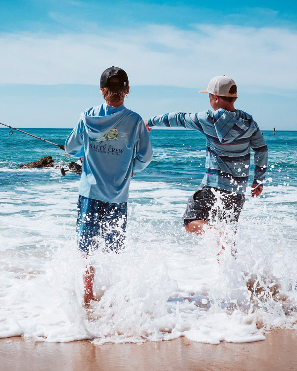Two boys stand in shallow ocean waves, with one holding a fishing rod. Both wear hooded shirts and caps, while water splashes around their legs under a bright blue sky.