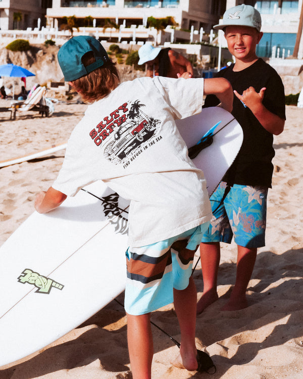 Two boys in swim trunks and hats stand on a sandy beach, smiling and holding surfboards. Beachgoers, umbrellas, and buildings are visible in the background.