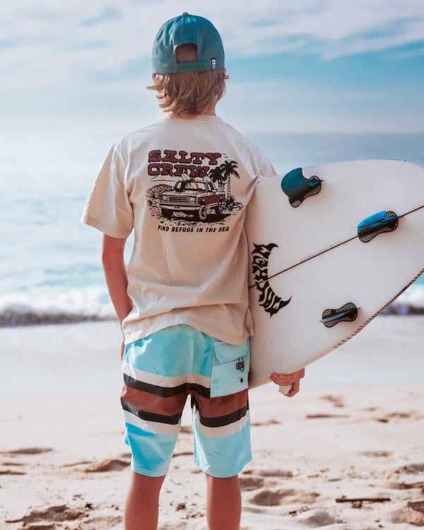 A person in a beige T-shirt, blue and brown board shorts, and a teal cap stands on the sandy beach holding a surfboard, facing the ocean under a partly cloudy sky.