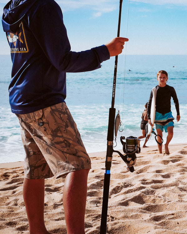 A person in a blue hoodie and camo shorts holds a fishing rod on a sandy beach, while two children with surfboards walk from the ocean in the background under a clear sky.