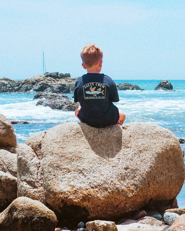 A young boy with light hair sits on a large rock by the ocean, facing away. He wears a navy “Salty Crew” t-shirt. The sea, rocky shoreline, and a distant sailboat are visible under a bright blue sky.